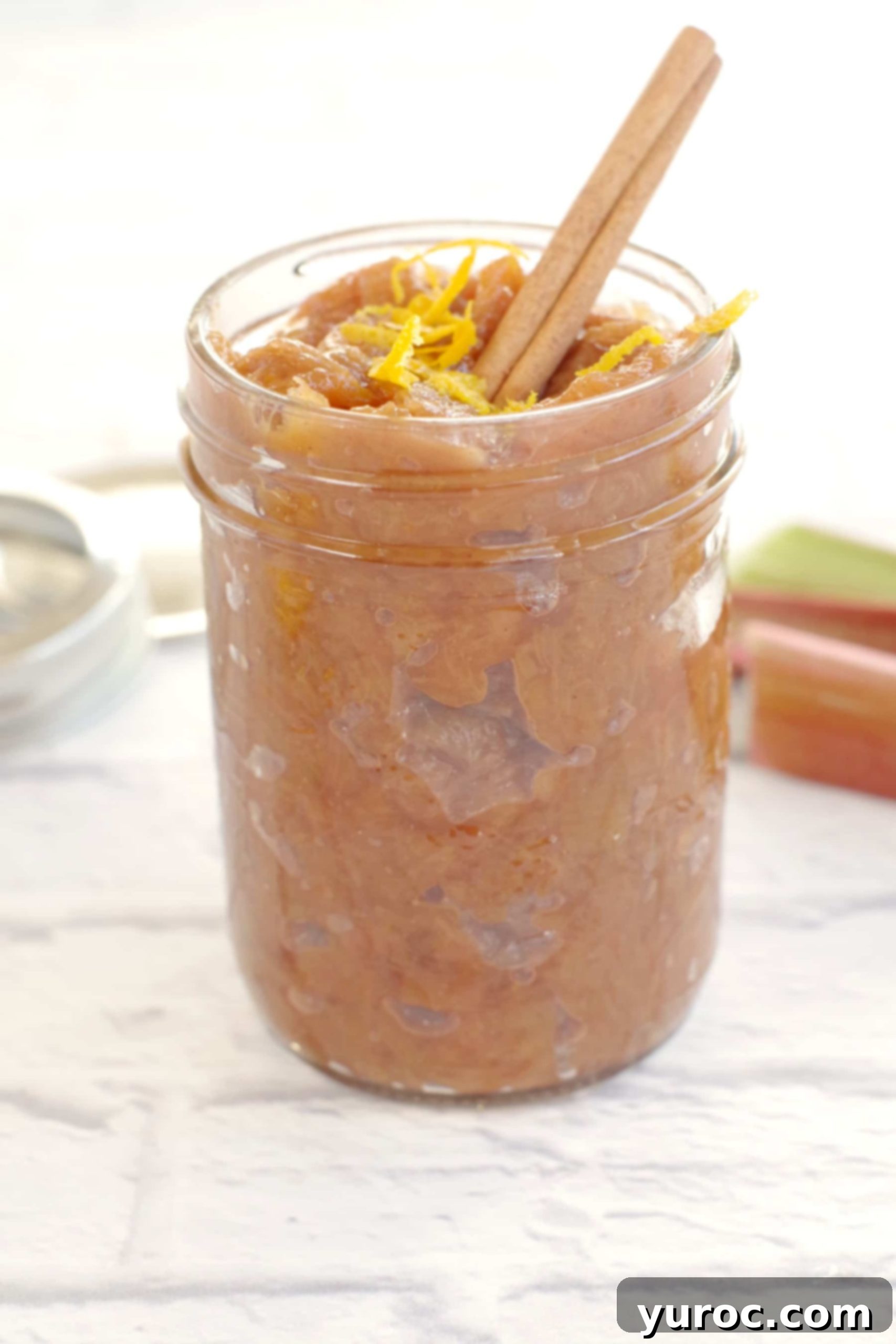 Close-up of homemade rhubarb compote in a clear glass jar, adorned with fresh orange zest and a cinnamon stick, ready to be enjoyed.