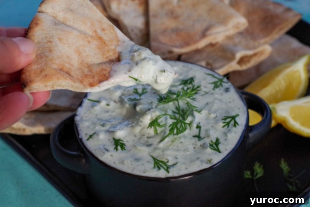 Grecian Baked Pasta Bites 5 triangle of pita bread being dipped into creamy tzatziki sauce in a black bowl on a black tray with pita bread in background and lemon wedges