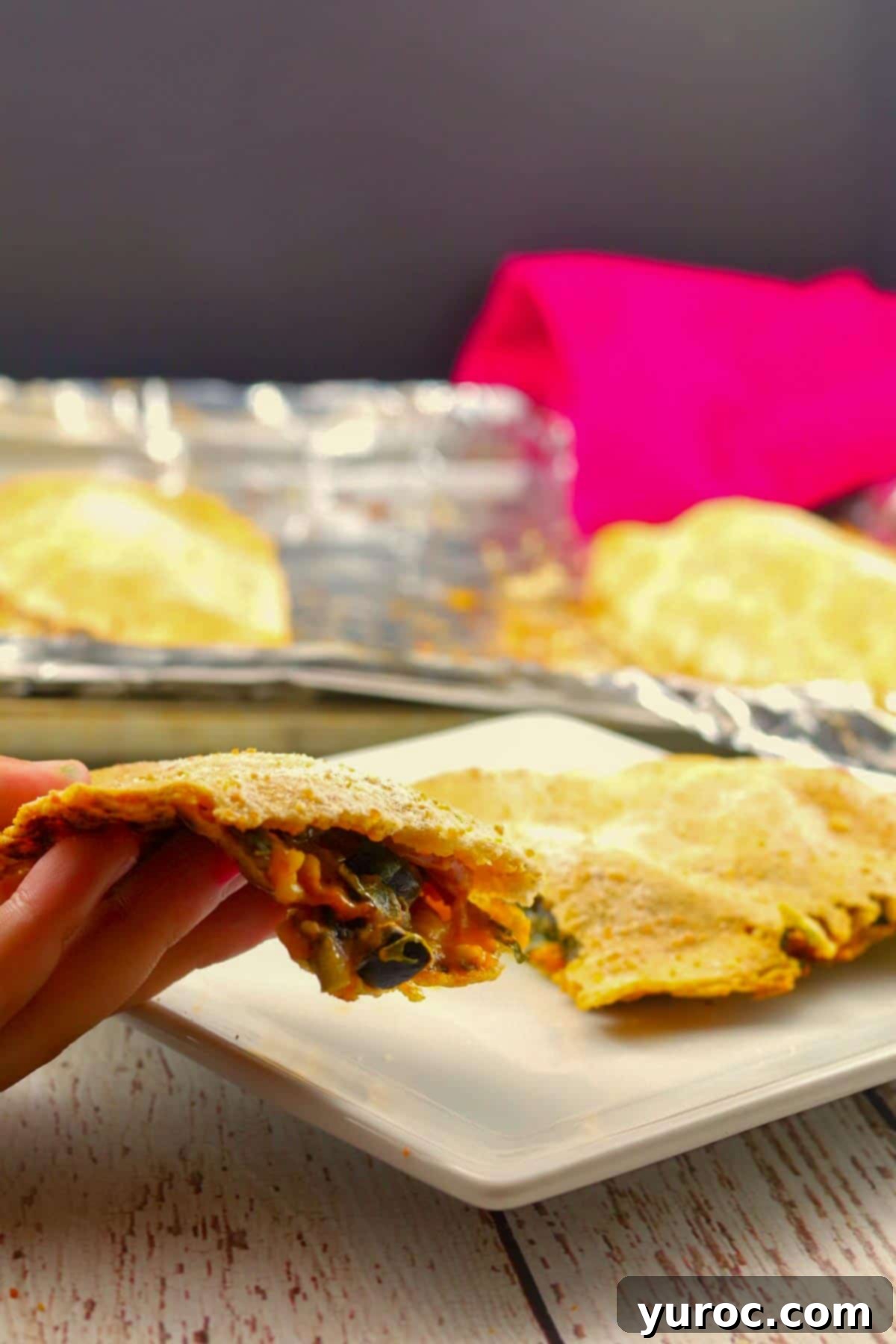 Wholesome Pita Pizza Pouches 5 A homemade hot pocket being held up by hand, with remaining hot pockets on a white plate and baking sheet in the background, showing off its golden crust.