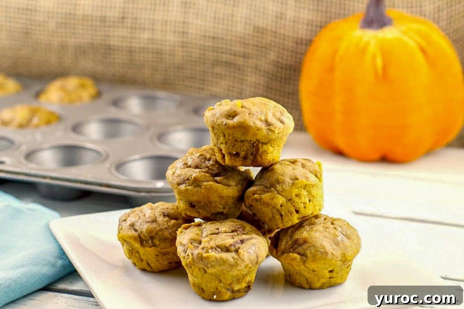 Weight Watchers mini pumpkin muffins stacked on a white plate with a mini muffin tin and pumpkin prop in the background