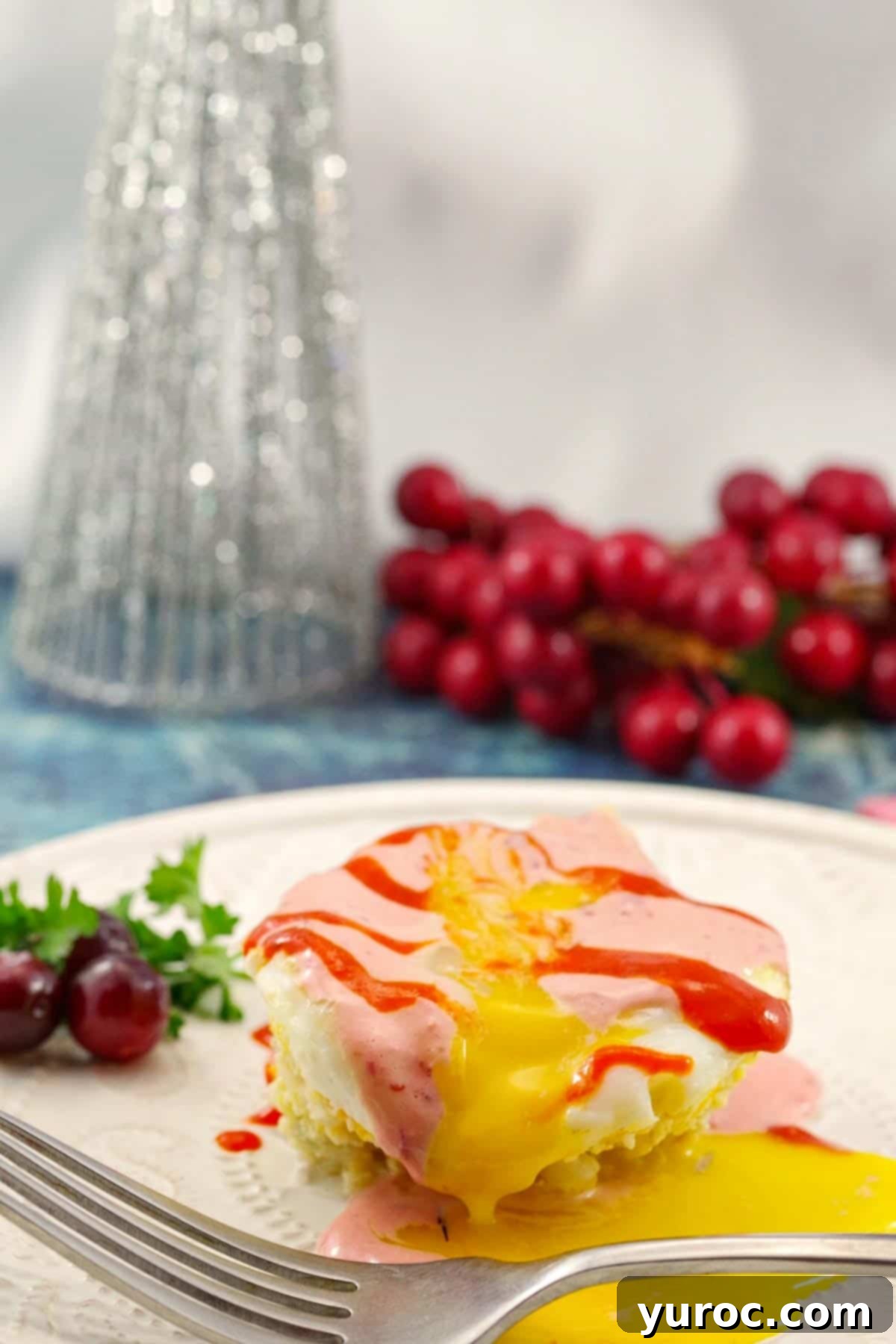 muffin tin eggs benedict on a white plate with cranberries and a Christmas tree decoration in the background