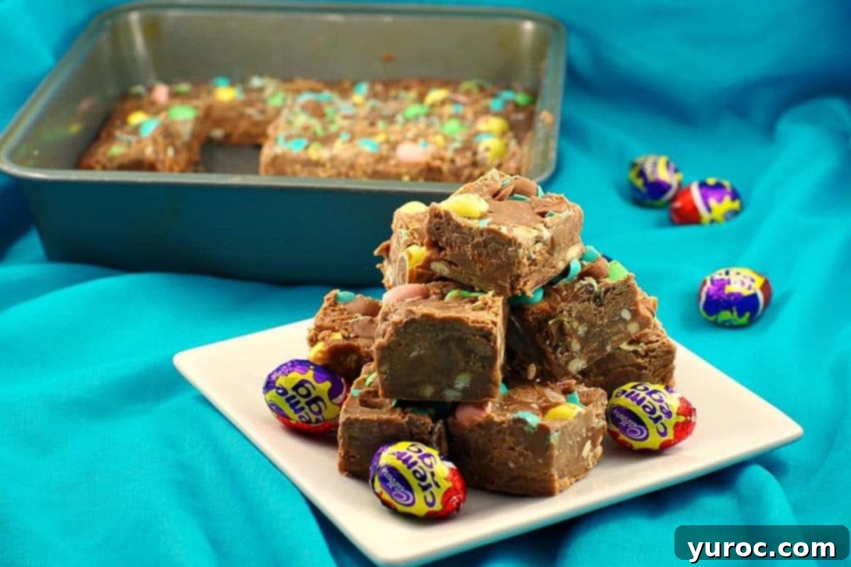 Microwave Easter Egg Fudge pieces stacked on a plate with a pan of more fudge in the background, showcasing its rich texture and colorful candy topping.
