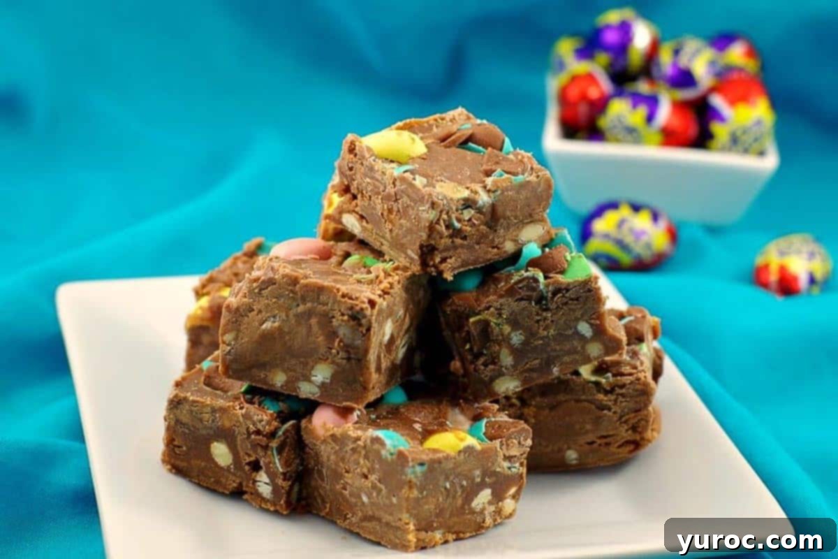 Microwave Easter Egg Fudge pieces neatly stacked on a white plate, with a bowl of colorful Cadbury Mini Easter Creme eggs in the background, highlighting the ingredients.