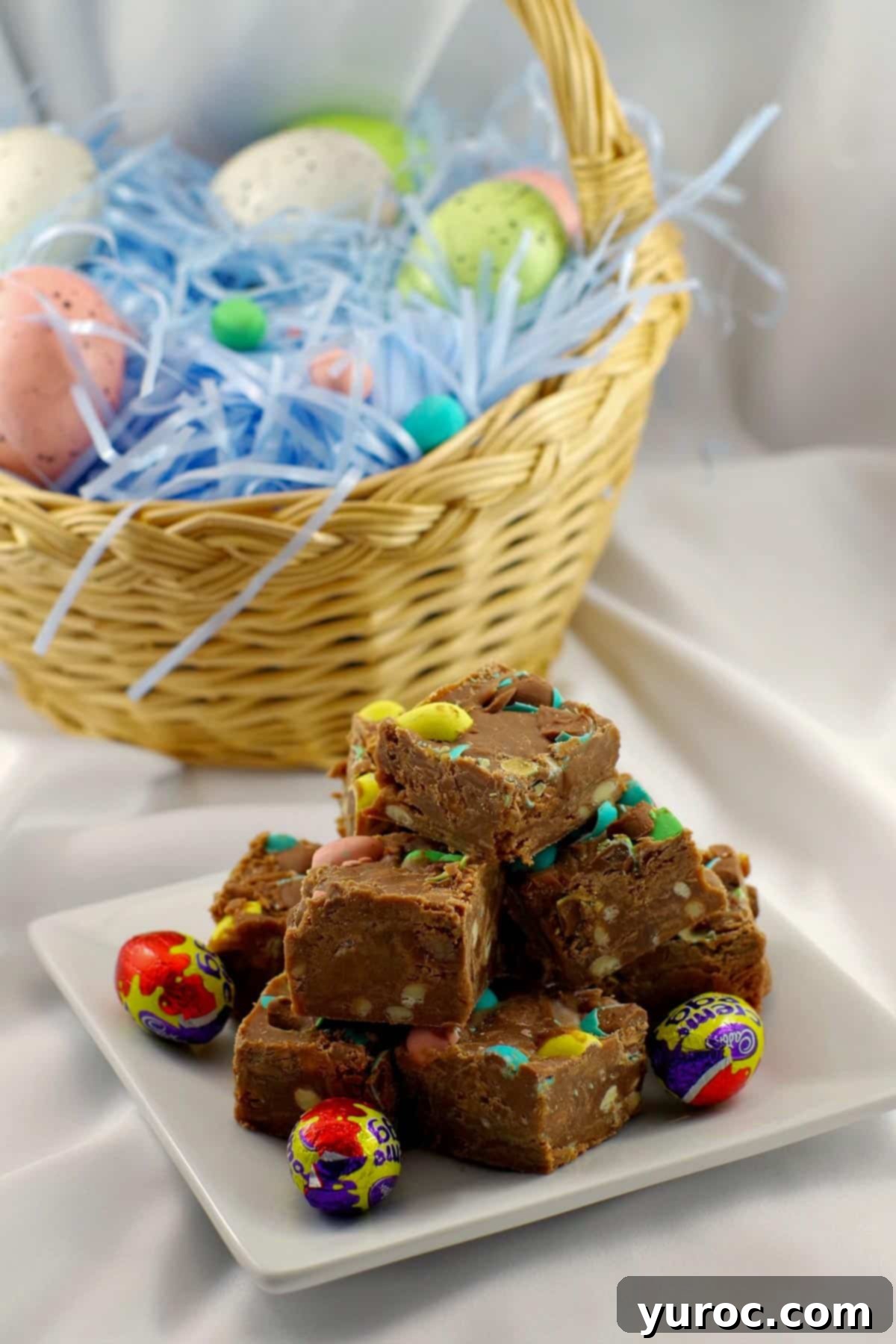 Easter Egg Fudge pieces stacked on a white plate, with a decorative Easter basket filled with eggs in the blurred background, emphasizing its festive appeal.