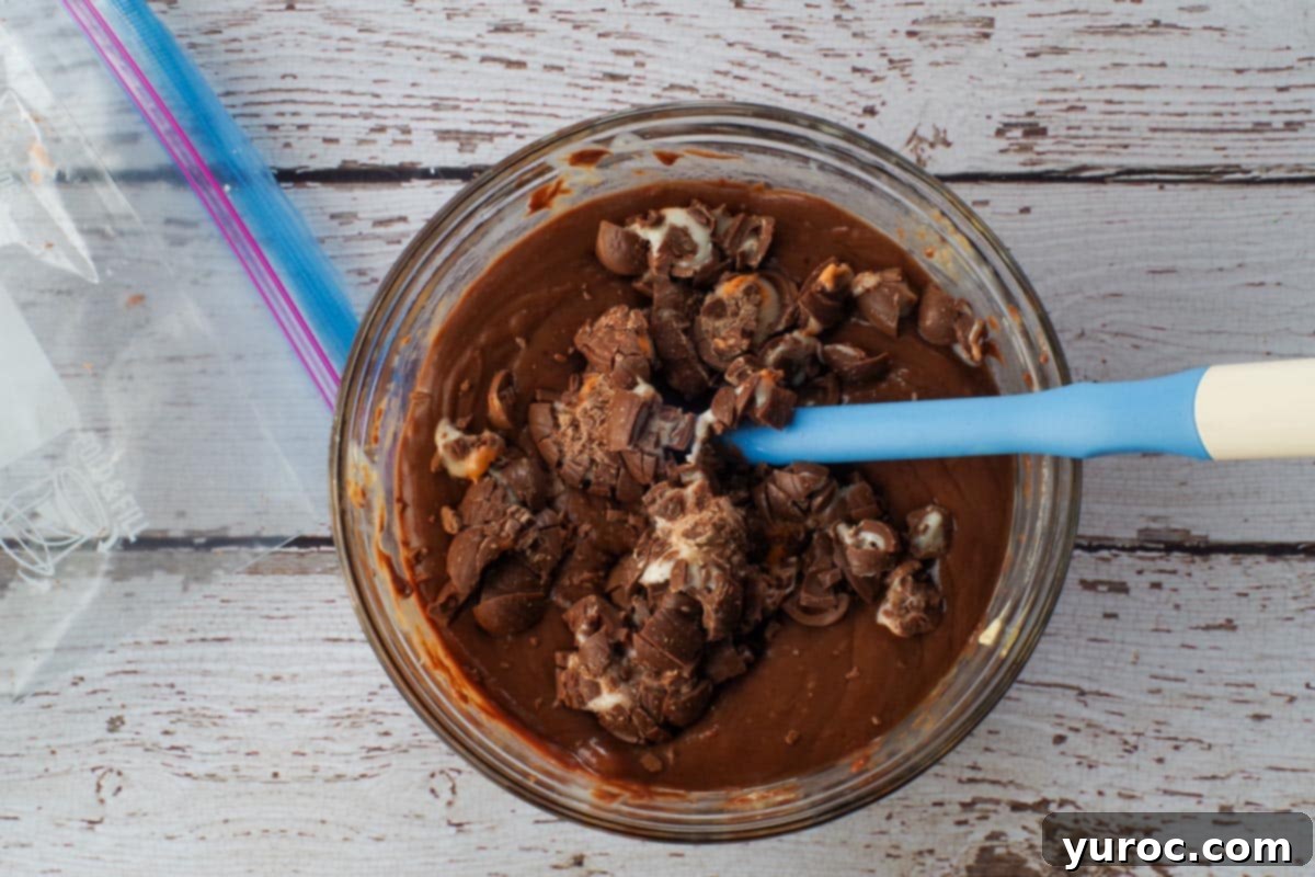 Crushed Cadbury Mini Creme Eggs being gently stirred into the melted chocolate fudge mixture in a glass bowl with a spatula, ensuring even distribution.