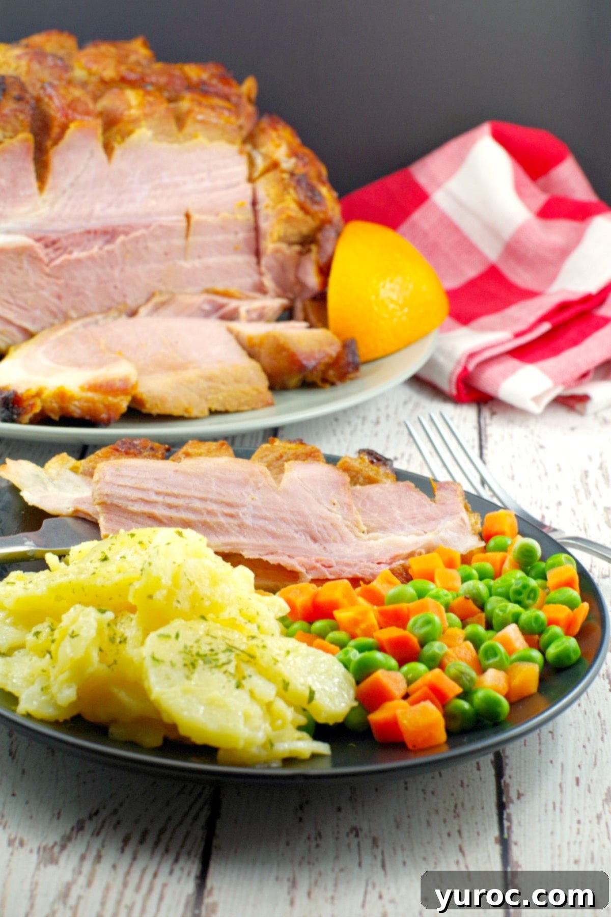 Sliced picnic ham on a plate with scalloped potatoes and veggies with whole ham in background