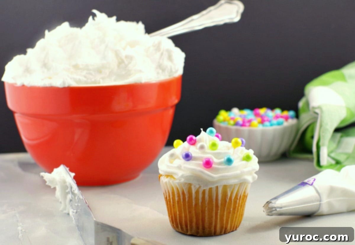 A beautifully frosted cupcake adorned with pearl sprinkles, a frosting spatula, and a decorating tip in the foreground. In the soft-focus background, an orange mixing bowl holds a generous amount of fluffy white frosting, alongside a small bowl of shimmering pearl sprinkles, highlighting the delicate elegance and essential tools for this unique buttercream recipe.