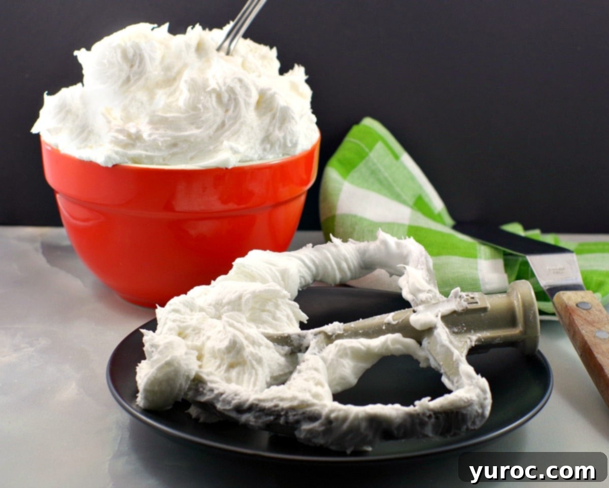 A stand mixer paddle attachment rests on a white plate, laden with a generous swirl of fluffy white icing, freshly prepared. In the background, an orange mixing bowl holds an abundance of the whipped frosting, showcasing its light and airy texture. This image highlights the luscious consistency and vibrant white color of the shortening-based buttercream, ready for decorating.