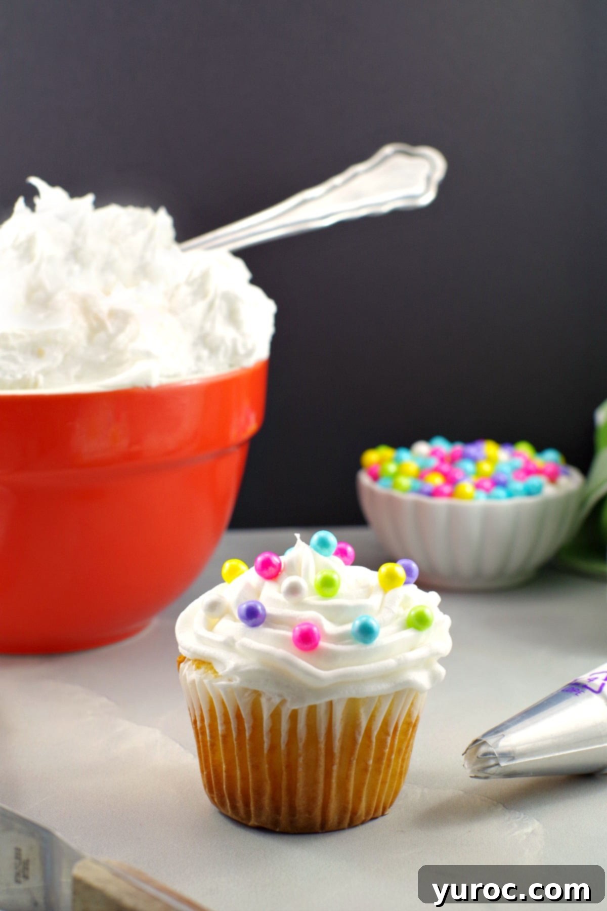 A charmingly frosted cupcake, adorned with delicate pearl sprinkles, is positioned in the foreground. Beside it lie a frosting spatula and a decorating tip, essential tools for creating beautiful designs. In the soft background, an inviting orange bowl overflows with more of the luxurious whipped frosting, accompanied by a small bowl of glistening pearl sprinkles, showcasing the delightful possibilities for cake decoration.