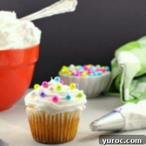 A cupcake, beautifully adorned with light, fluffy frosting and delicate pearl sprinkles, is positioned in the foreground. A frosting spatula and a decorating tip lie beside it, hinting at the creative process. In the background, a vibrant orange bowl holds a generous amount of whipped frosting, accompanied by a small bowl of pearl sprinkles, showcasing the delightful potential of this shortening-based buttercream recipe.