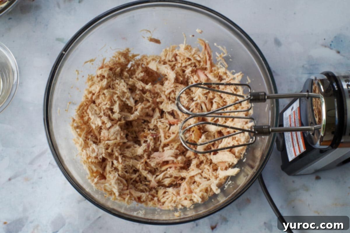 Shredded chicken in large glass bowl with beaters above bowl.