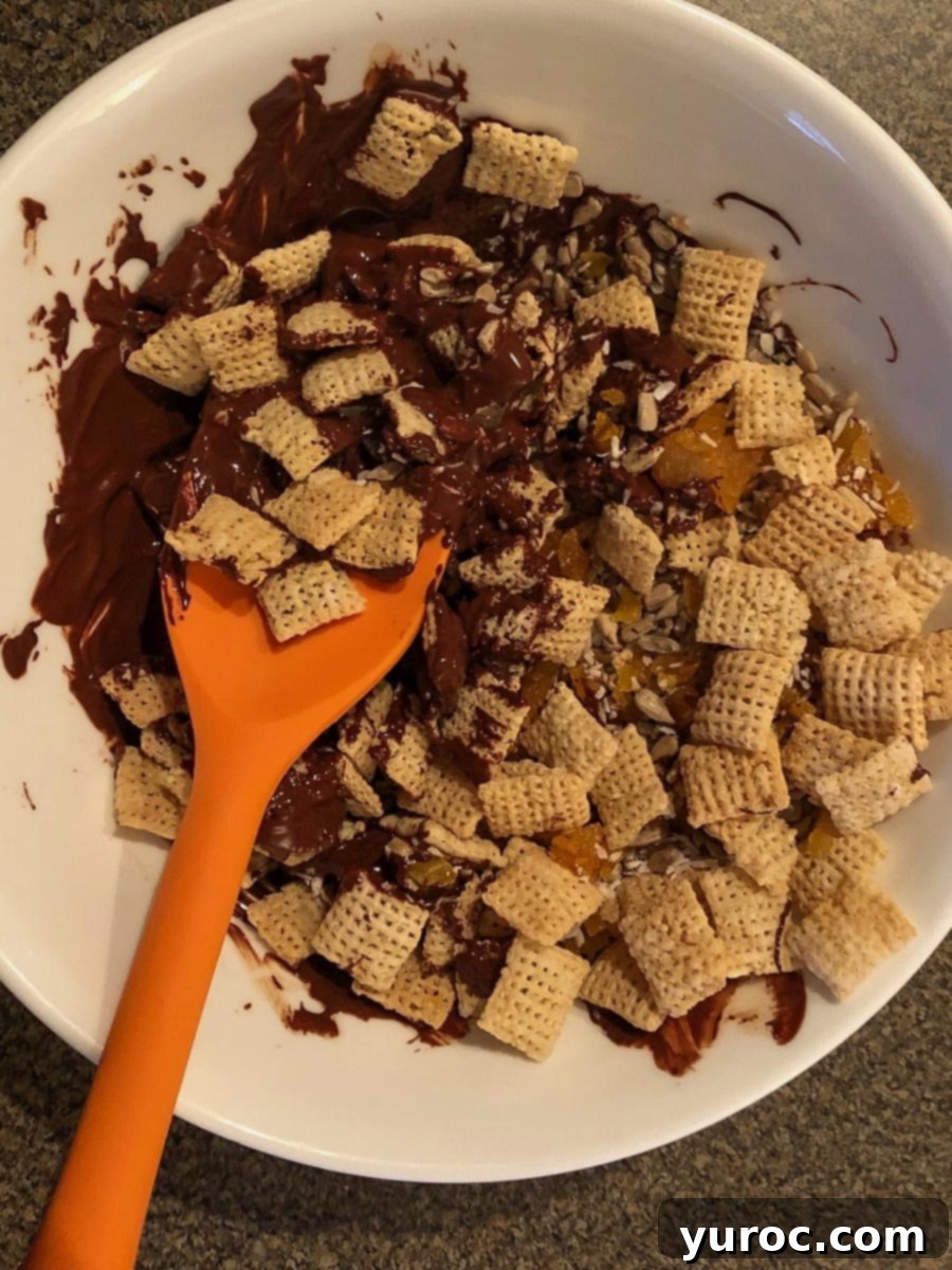 Cereal mixture being added to melted chocolate in a white bowl with orange spatula.