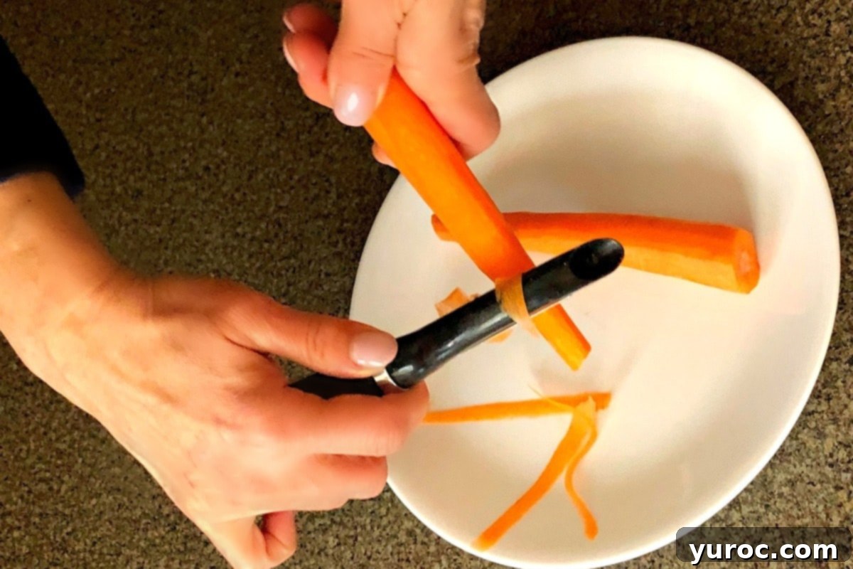 A hand holding a vegetable peeler is creating thin, delicate ribbons from a fresh carrot over a white surface, demonstrating the technique for making an elegant garnish.