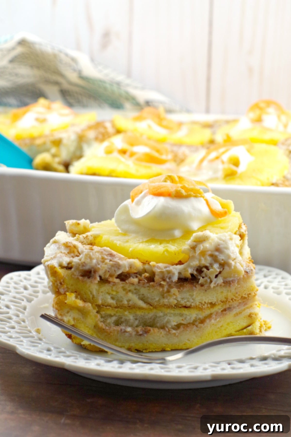 A perfectly portioned slice of Carrot Cake French Toast Casserole sits in the foreground, showcasing its rich, custardy layers with pineapple and walnuts. In the background, the full casserole, beautifully glazed and garnished, awaits, emphasizing its appeal as a breakfast or brunch item.