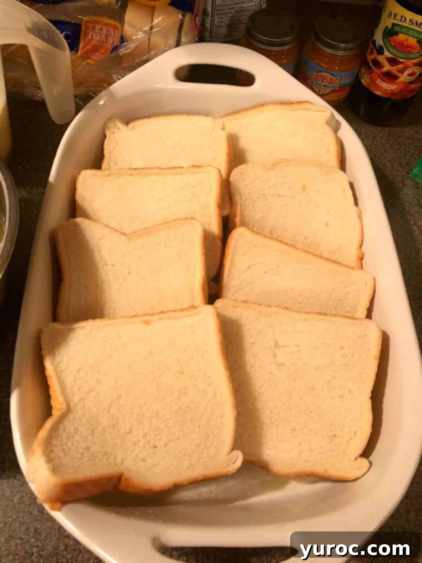 Slices of thick French bread are arranged neatly and slightly overlapping in two rows within a white rectangular casserole dish, ready to be soaked for a French toast casserole.
