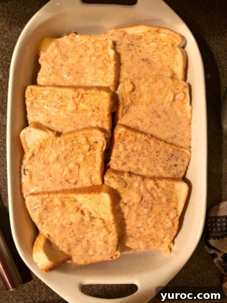A rich, creamy French toast mixture, speckled with pineapple and carrots, is being poured from a large bowl over a layer of bread slices in a white casserole dish, ensuring even saturation.