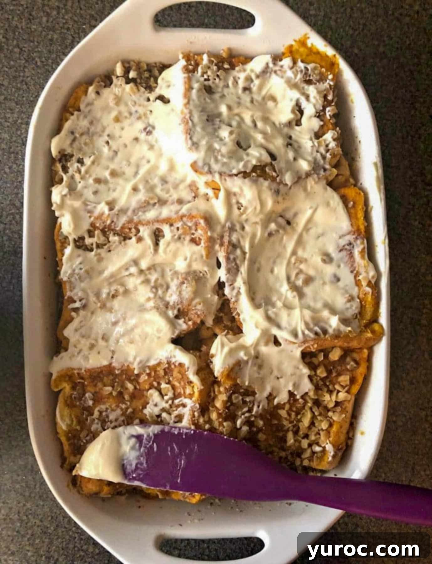 A spatula is carefully spreading a thick, white cream cheese glaze over the warm, golden-brown surface of a baked Carrot Cake French Toast Casserole in a white baking dish.