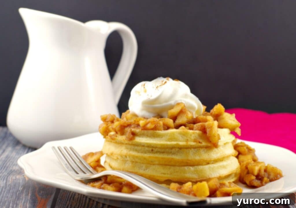 Warm Apple Cinnamon Topping 6 Crispy waffles generously adorned with warm apple cinnamon topping, elegantly presented on a white plate with a white syrup container visible in the soft background.
