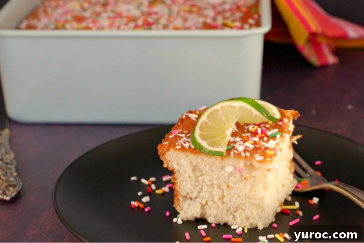 A slice of Coconut Tres Leche Cake adorned with sprinkles and a lime twist, served on a black plate. An 8x8 baking pan containing more of the cake is visible in the soft-focus background.