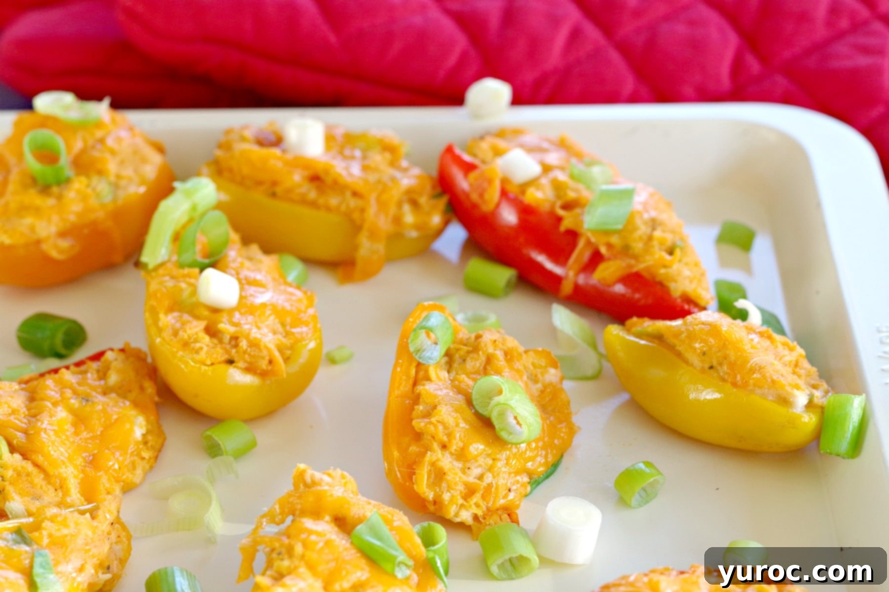 Zesty Buffalo Chicken Pepper Poppers 2 Buffalo chicken stuffed mini peppers on an off-white baking sheet with red oven mitts in the background, ready to be baked.