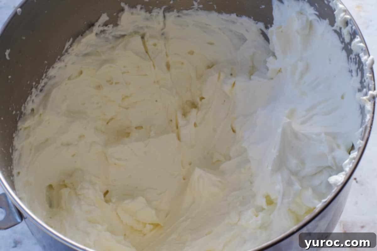 The white chocolate frosting, now transformed into a light and fluffy whipped ganache, shows perfectly stiff peaks in the bowl of a stand mixer after being beaten.