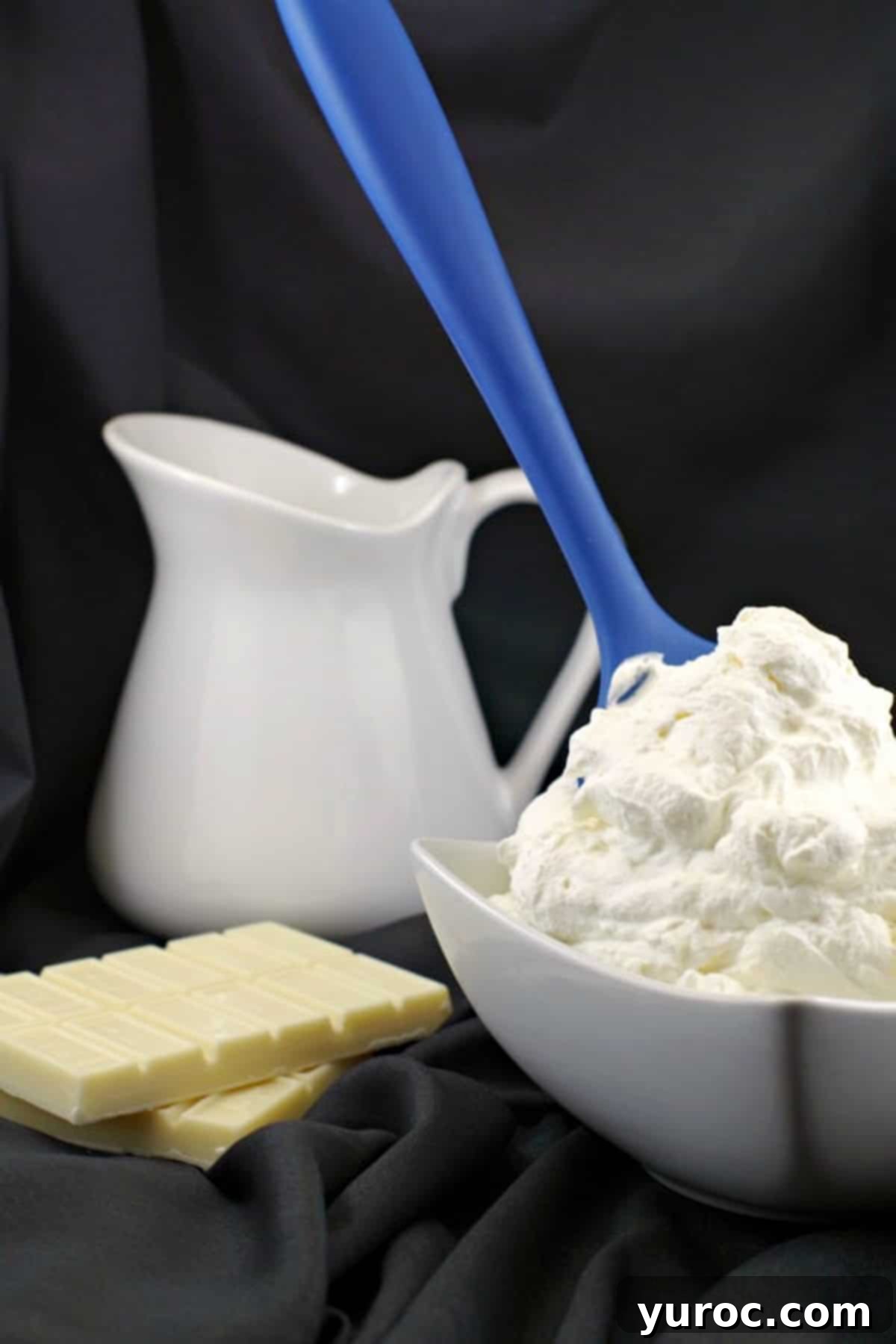 A large, inviting bowl of white chocolate whipped cream frosting, with a blue spatula handle protruding from it, and a white cream container blurred in the background, showcasing its delightful texture.