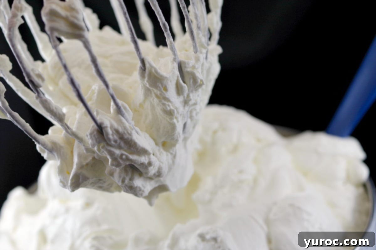 A whisk attachment from an electric mixer being lifted from a bowl of perfectly whipped white chocolate cream frosting, showing its fluffy texture.