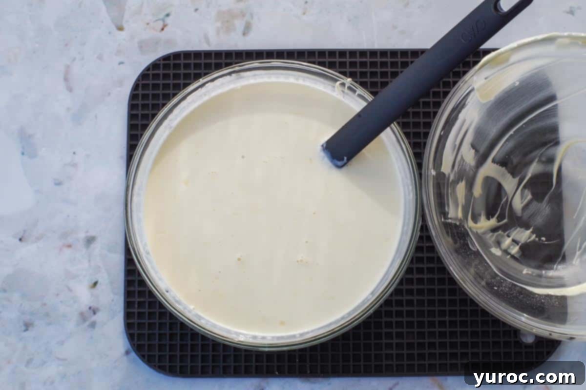 Warm whipping cream and melted white chocolate expertly combined and stirred together in a clear glass bowl, forming a smooth mixture.
