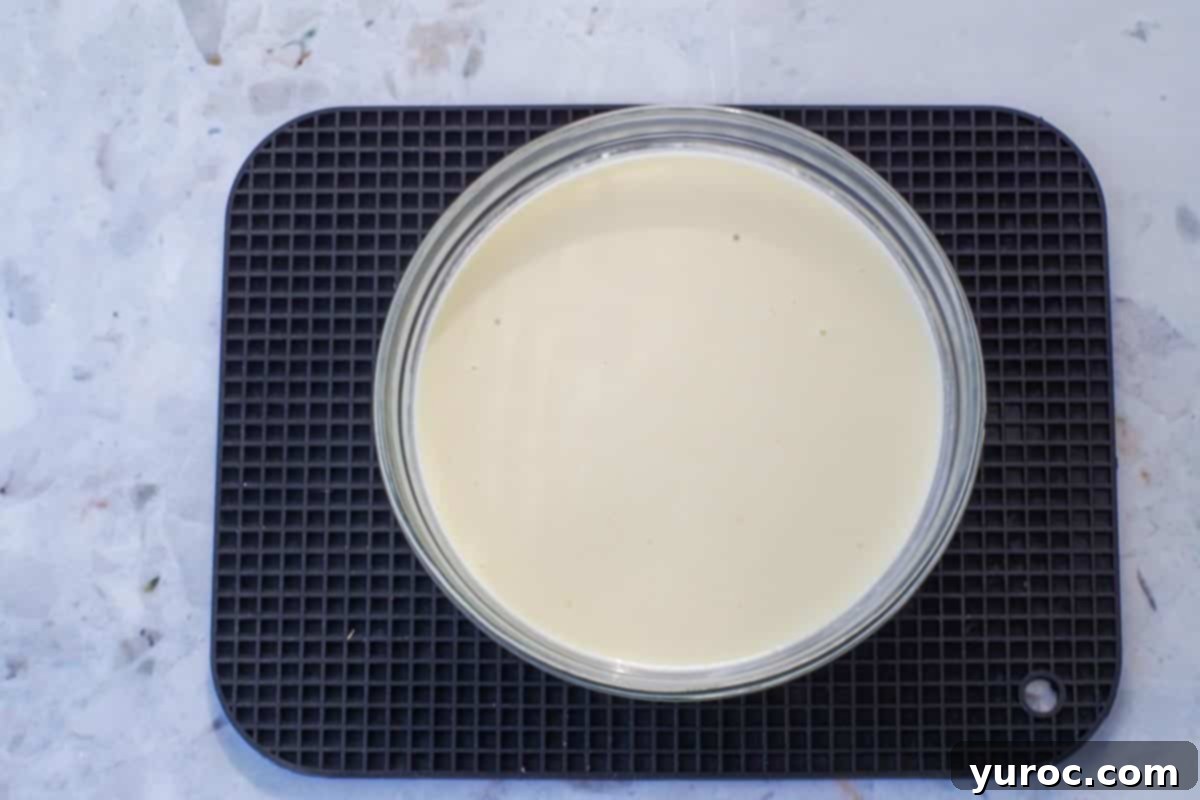 A glass bowl containing the smooth white chocolate and whipping cream mixture, resting on a rectangular black trivet to cool at room temperature.