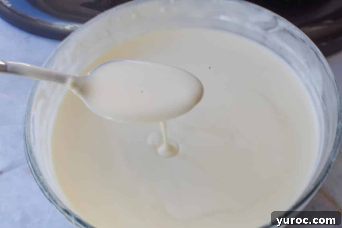 A spoon lifting a portion of the chilled white chocolate whipping cream mixture, demonstrating its thick, gravy-like consistency before whipping.