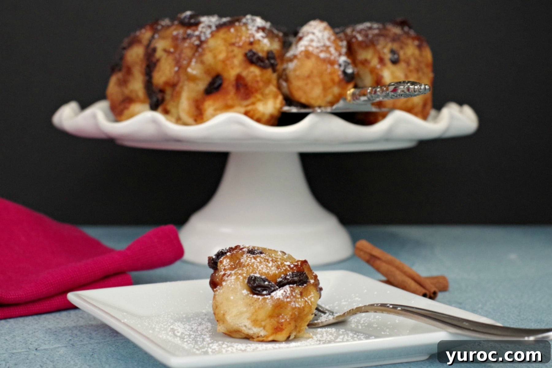 Overnight Caramel Cinnamon Buns 2 Overnight Cinnamon Sticky Buns on a white cake platter with bun on white plate in front, showcasing their golden-brown exterior and sticky glaze.