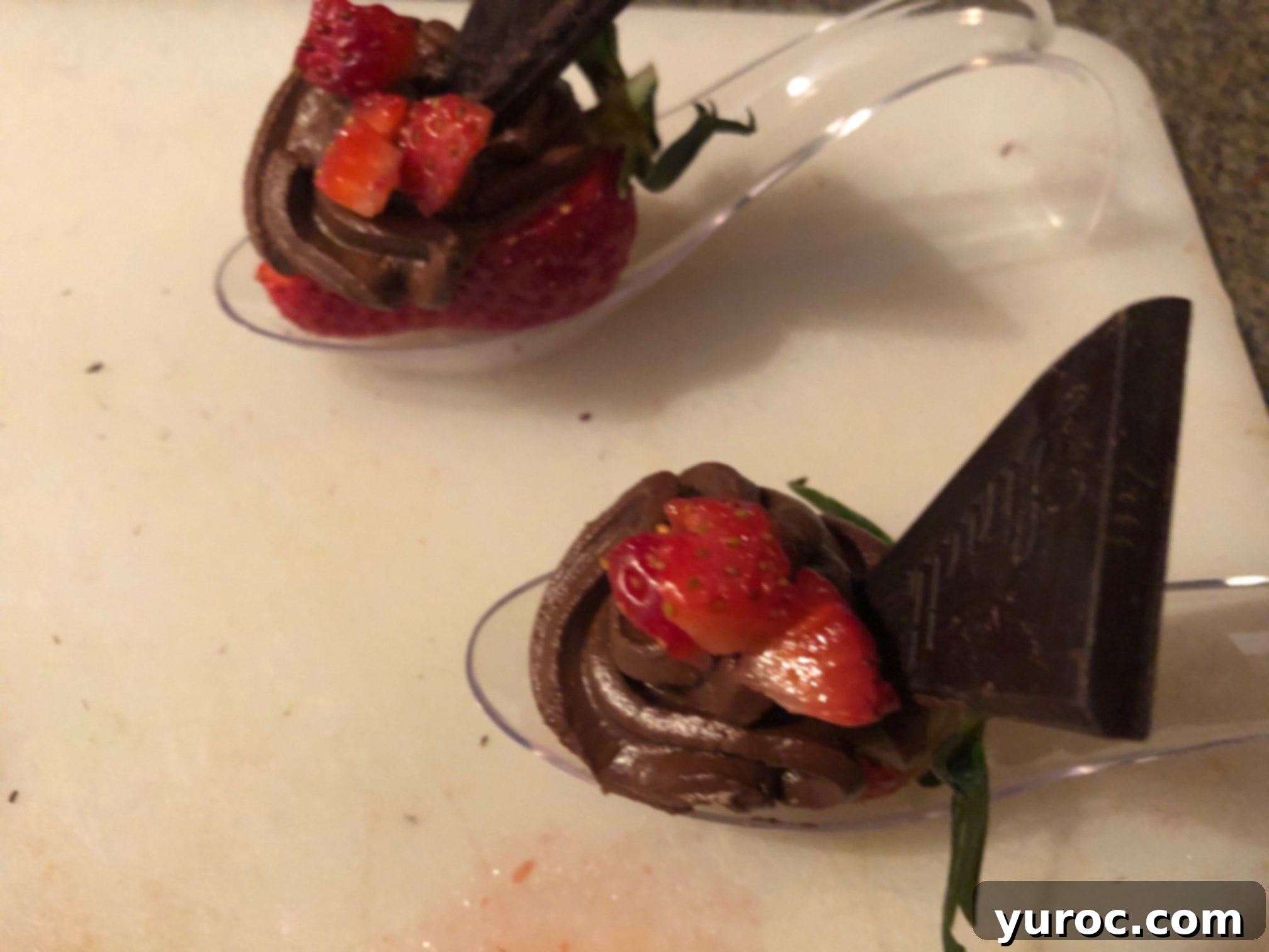 Finely chopped strawberries sprinkled on top of the chocolate avocado mousse, positioned in front of the dark chocolate piece on the spoon dessert.