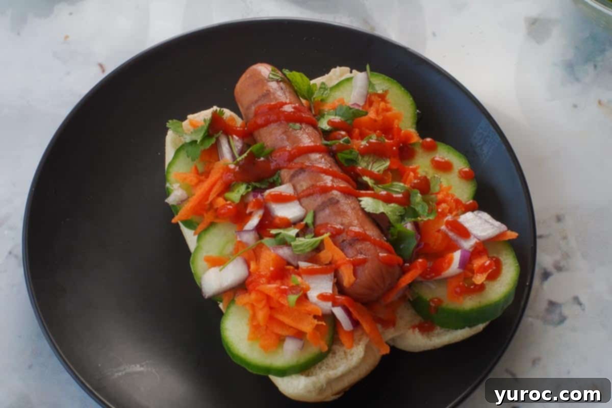 Banh Mi Turkey Hot Dog Twist 7 Veggies with turkey dog in a bun, with rice vinegar and sriracha sauce drizzled on it, presented on a black plate.