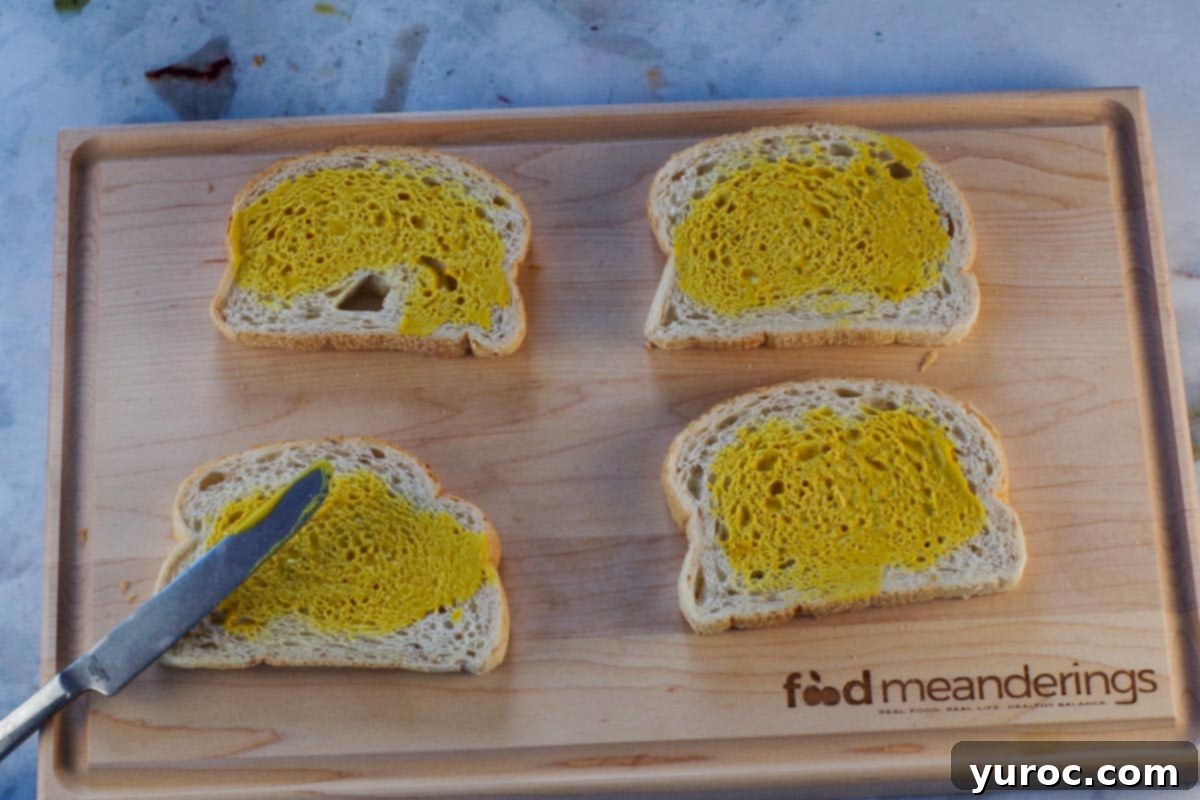 Mustard being spread on 4 pieces of bread on cutting board.