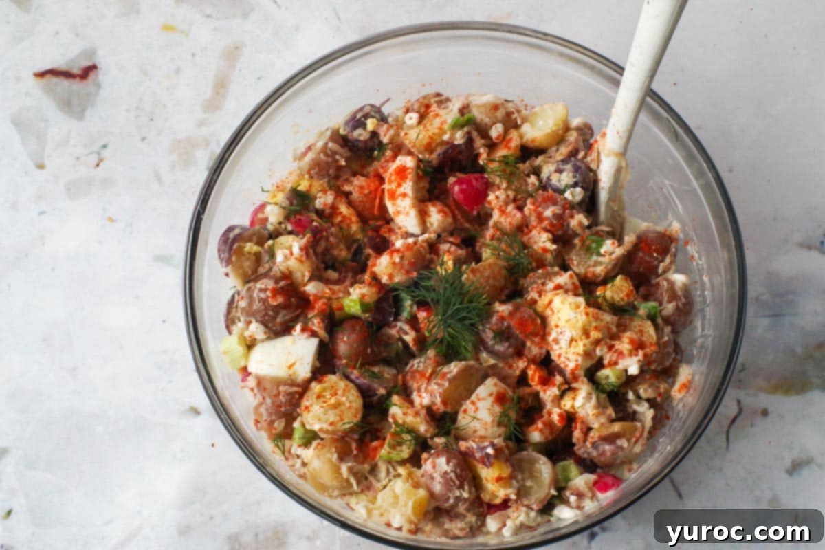 Signature Pierogi Potato Salad 14 Garnishes added to potato salad in large glass bowl with spoon.
