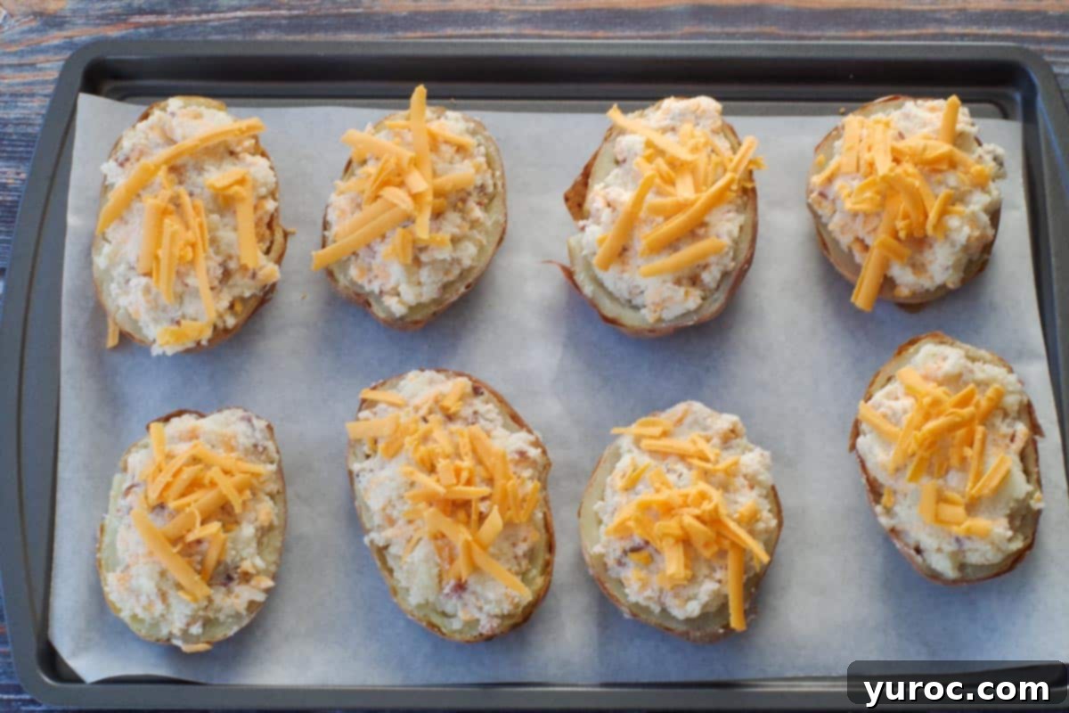 Prepared twice baked Caesar potatoes on a parchment-lined baking sheet, with the reserved cheddar cheese sprinkled over the tops, ready to be baked to golden perfection.