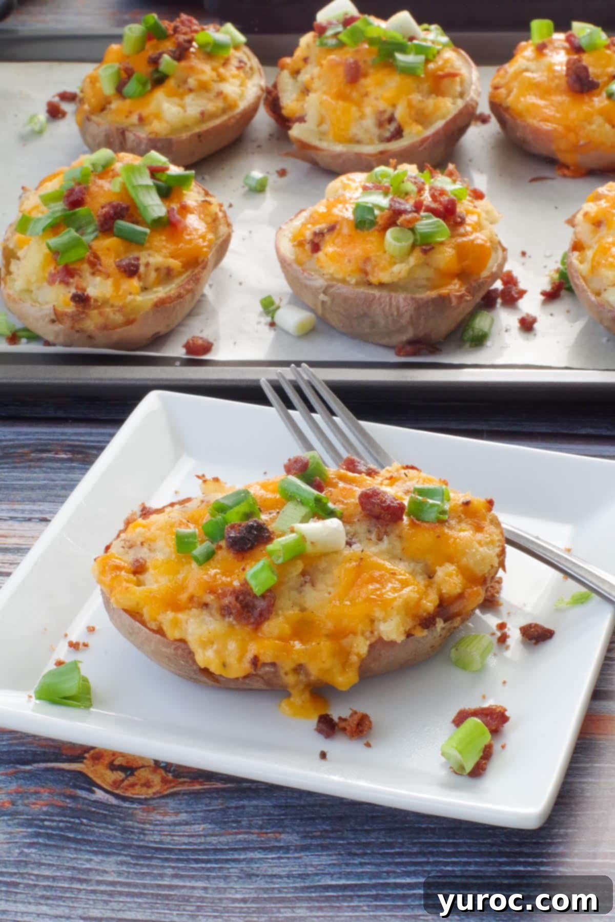 A freshly reheated Caesar twice baked potato on a white plate with a fork, ready to be eaten. In the blurred background, a sheet pan with more potatoes suggests a larger batch ready for enjoyment.