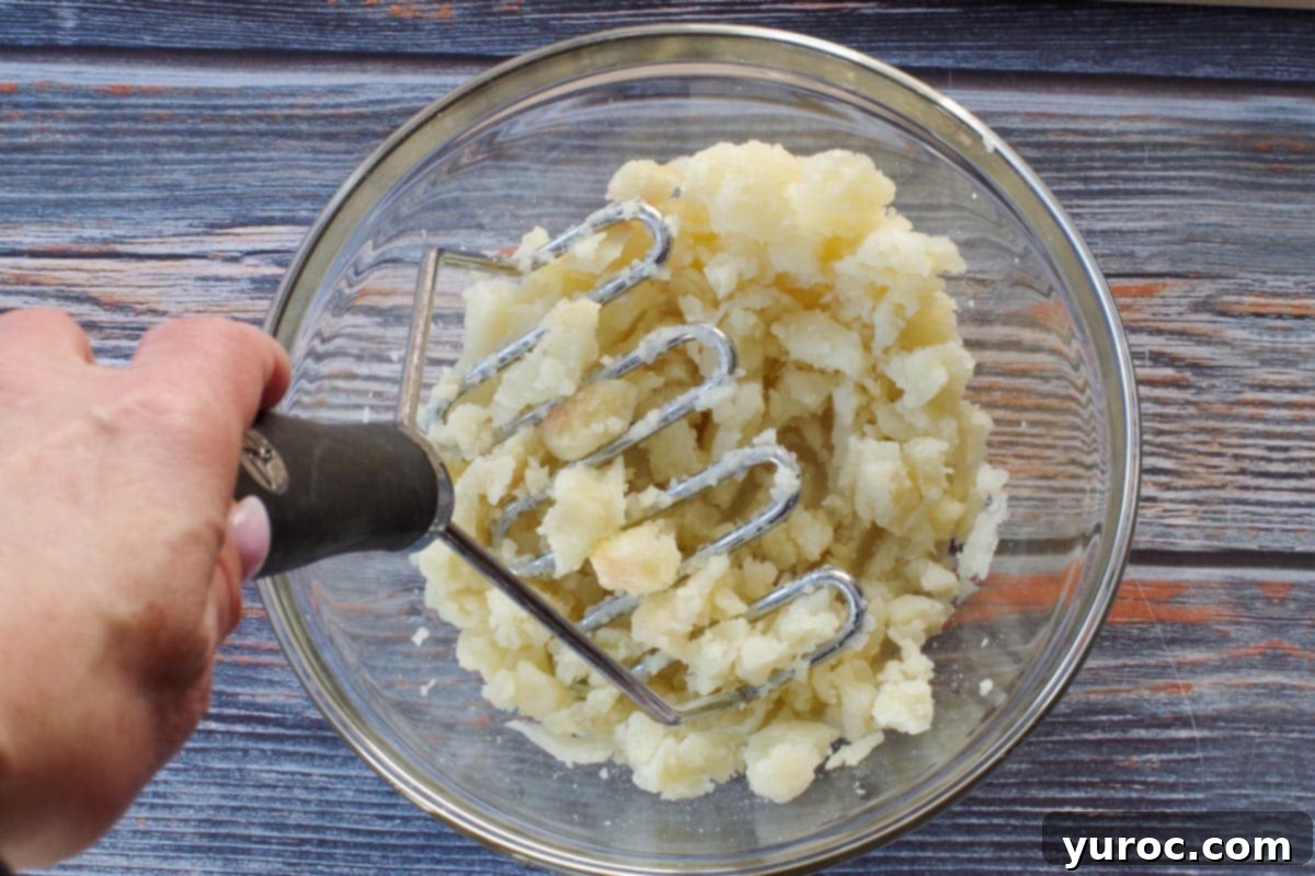 Fluffy potato flesh being thoroughly mashed with a potato masher in a clear glass bowl, ensuring a smooth and creamy base for the filling.