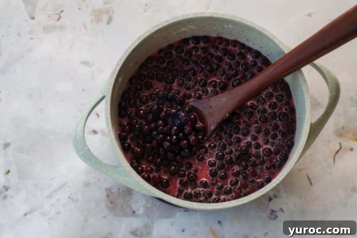 Saskatoon Berry Pie: Mastering the Filling and Sauce 5 Saskatoon berry mixture, in a blue pot, being stirred with a wooden spoon, as the berries are starting to defrost.