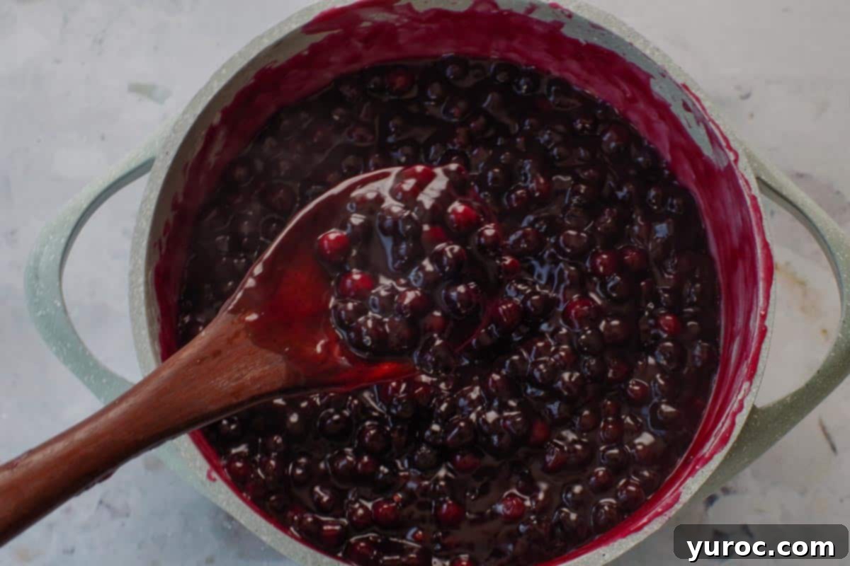 Saskatoon Berry Pie: Mastering the Filling and Sauce 8 Saskatoon berry sauce being held up on a wooden spoon to show thickeness (like jam) over blue pot.