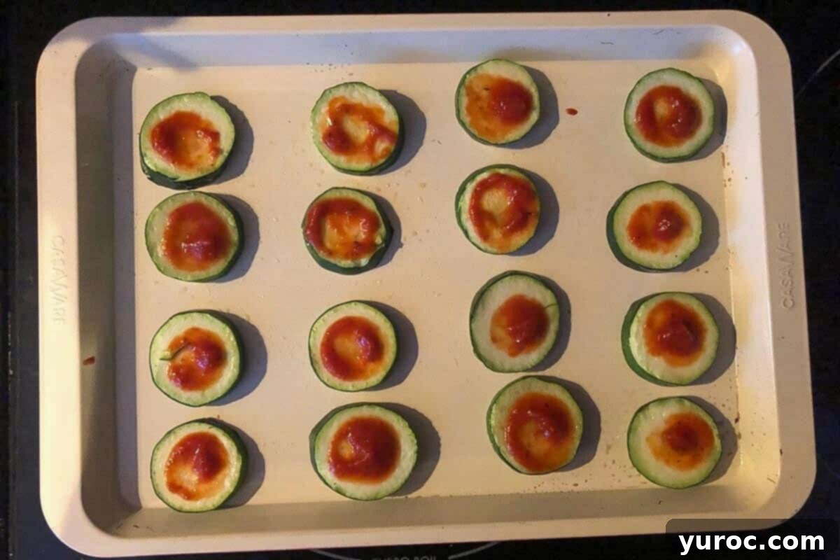 Zucchini rounds on a baking sheet, each topped with a small dollop of pizza sauce.