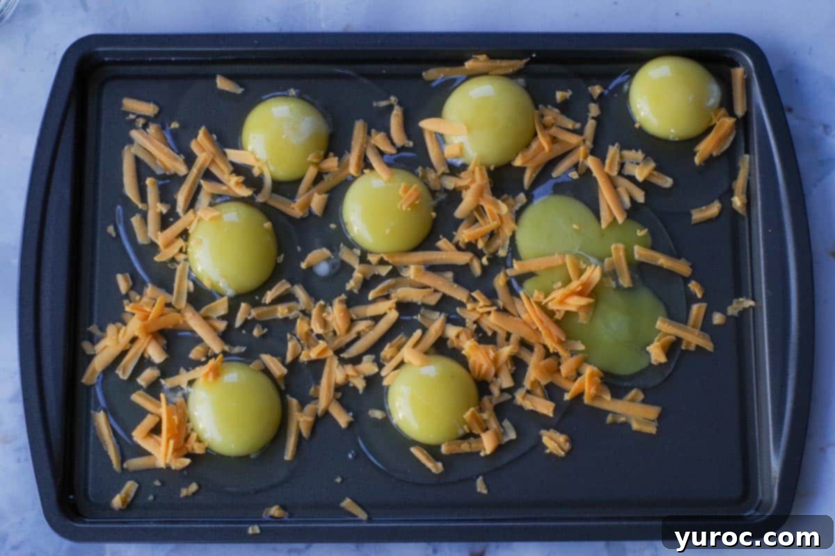 Raw eggs on a sheet pan with shredded cheddar cheese sprinkled over them.