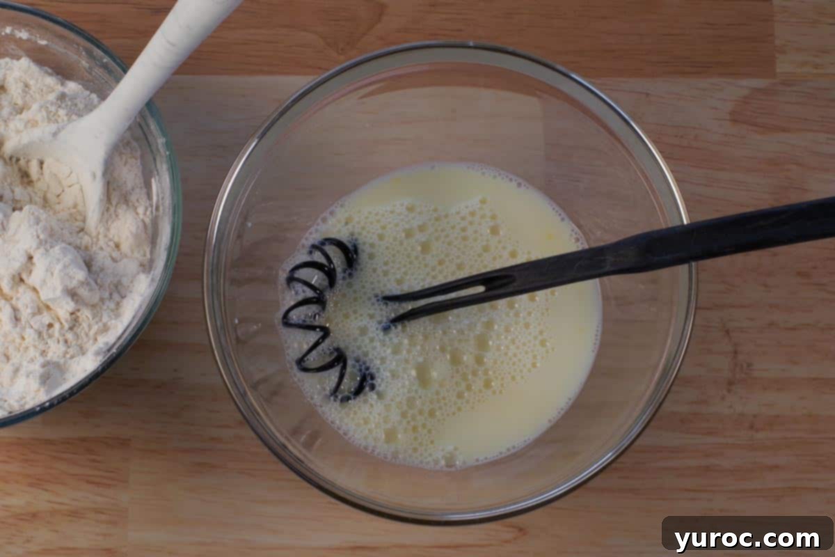 Egg and milk whisked together in a separate glass bowl with a black whisk, forming the wet ingredients for the biscuit dough.