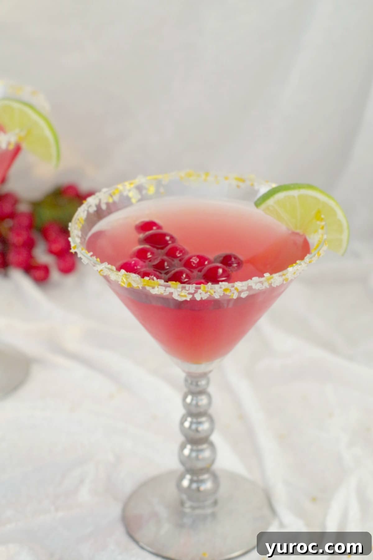 Cranberry Cosmo Mocktail with cranberries floating in drink, wedge of lime and sparkly gold sugar with cranberries in the background, on white material