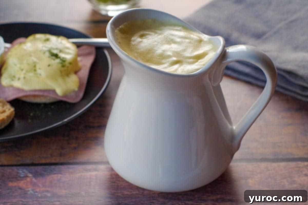 Healthy Hollandaise sauce in a large white creamer with plate of eggs benedict in background and grey linen