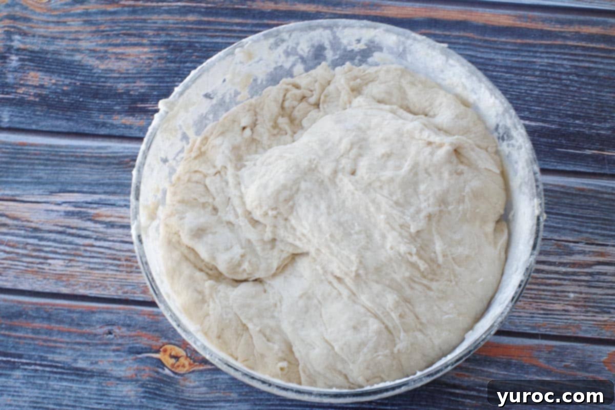 Additional flour being gradually added to the sticky dough mixture in a glass bowl