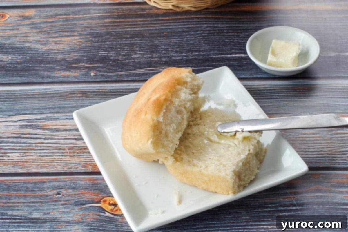 A Granny Bun split in half, with butter spread on one side, presented on a square white plate. A dish of butter sits in the background.