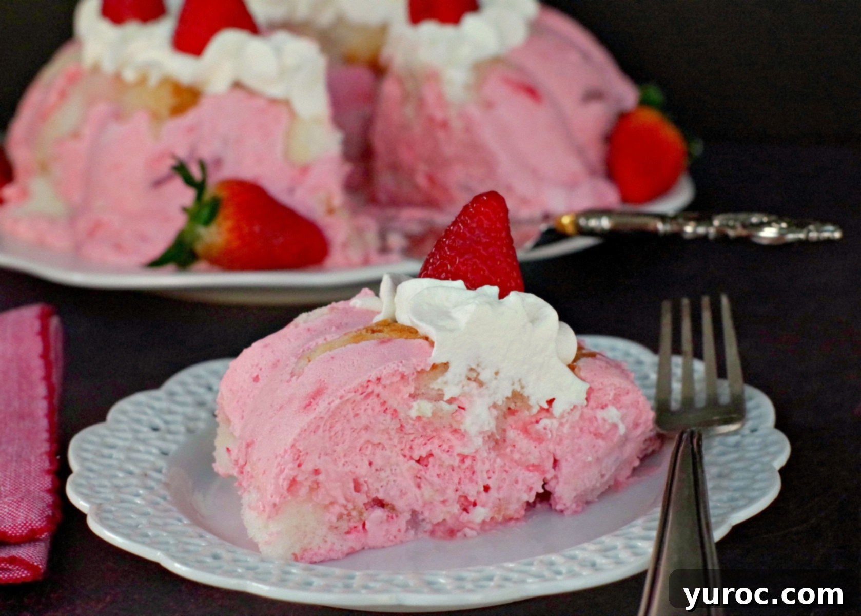 A slice of vibrant strawberry jello angel food cake on a white plate, with the whole cake blurred in the background