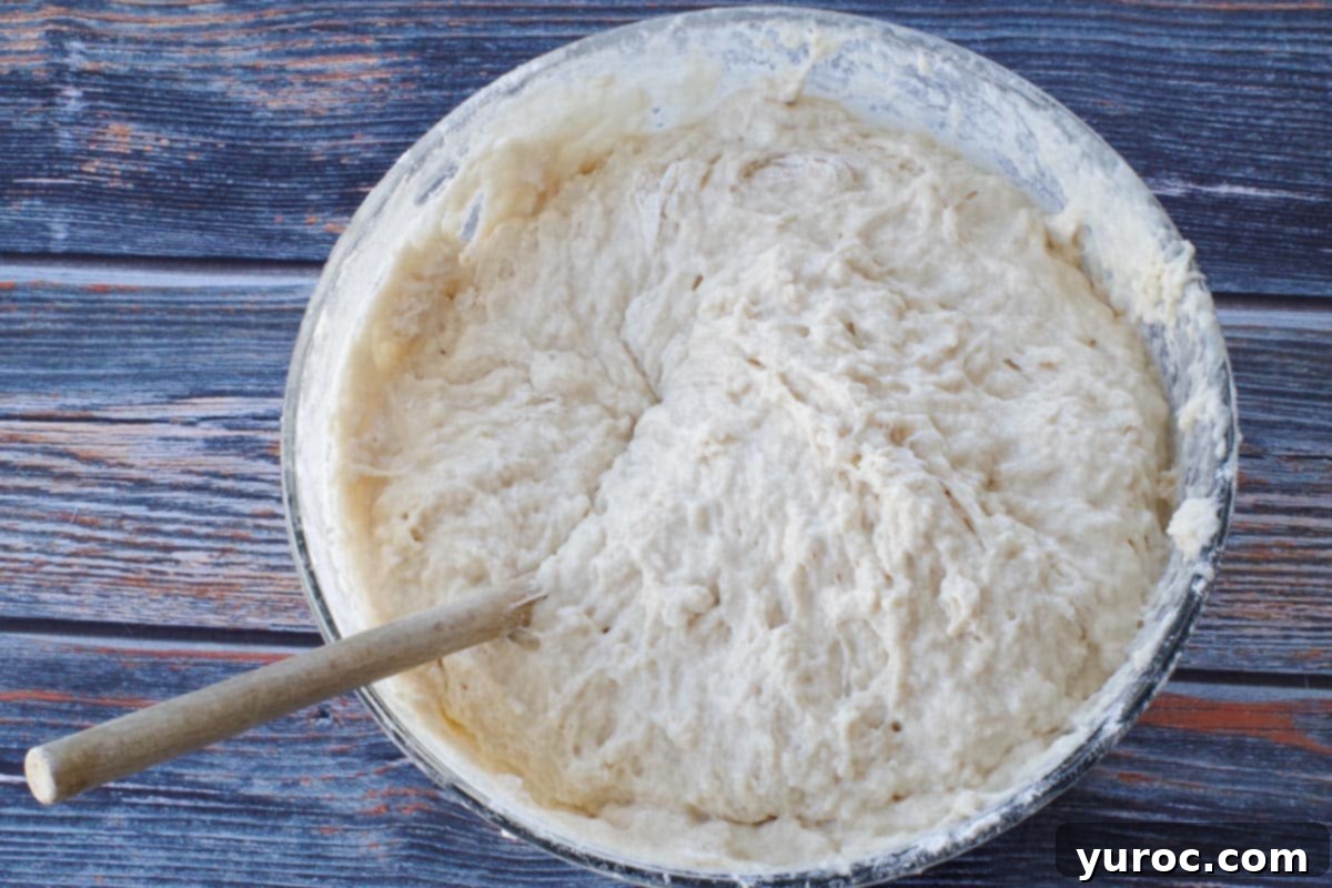 More flour being incorporated into the dough mixture in a glass bowl with a wooden spoon