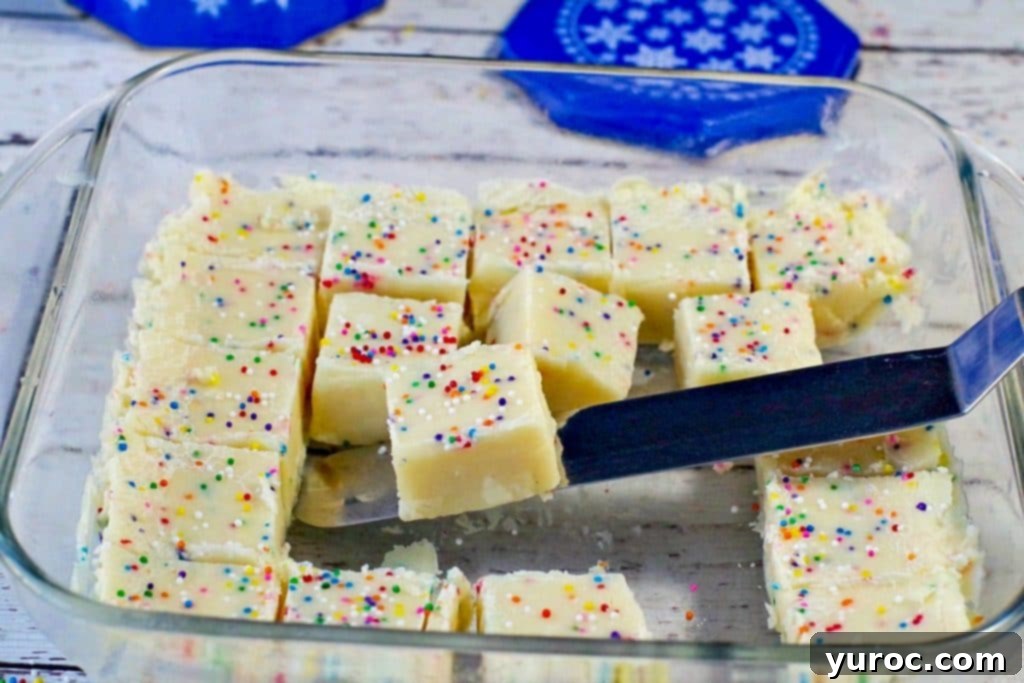 Easy Sugar Cookie Fudge 2 Delightful squares of sugar cookie fudge neatly arranged in a clear glass container, with a spatula gently lifting one piece, showcasing its creamy texture.